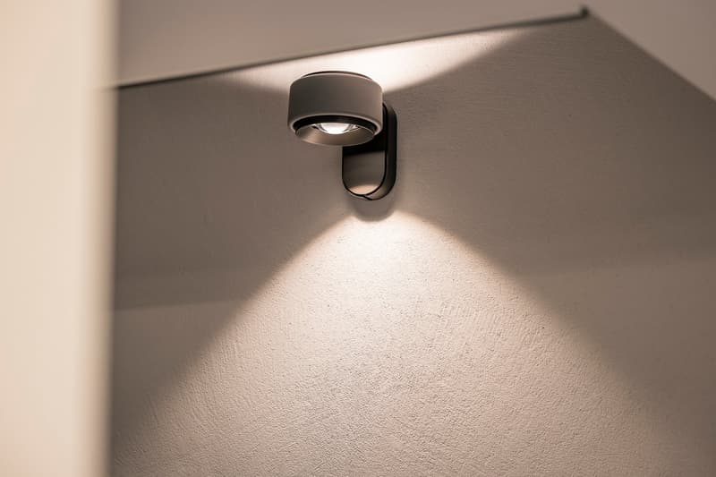 A modern, black wall-mounted light fixture with a round, focused beam of light illuminating a plain, white wall.