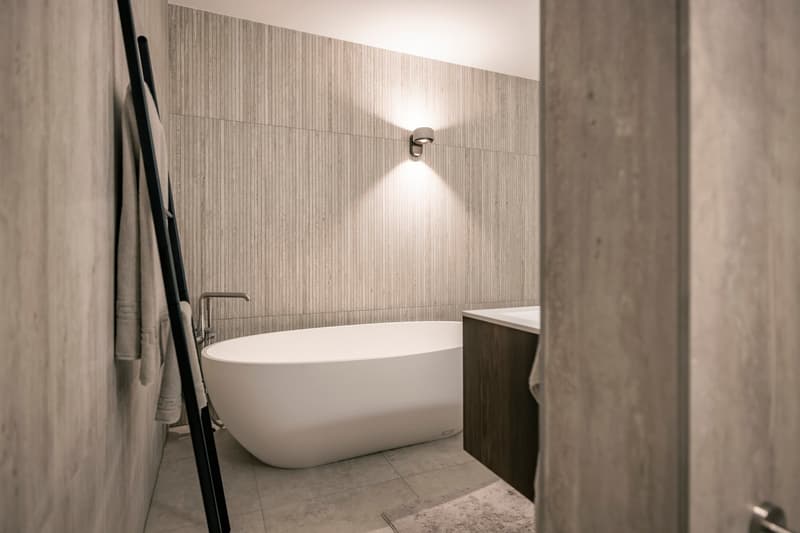 Spacious bathroom with a freestanding bathtub, wooden vanity, and tiled walls with vertical striped pattern. The room is well-lit by a wall sconce.