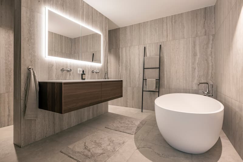 Spacious bathroom with gray marble-like tile walls, a freestanding bathtub, a floating vanity with drawers, and a towel rack. The bathroom also features a large backlit mirror and minimalist fixtures.