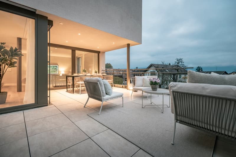 Spacious outdoor living area with a covered patio, comfortable seating, and a dining table. The space features large windows that provide a view of the surrounding landscape. The patio is furnished with modern, minimalist furniture and decor, creating a c