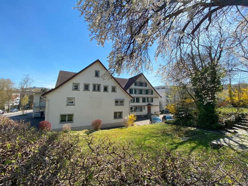 House with a brown roof, large windows, multiple floors, white exterior, garden area, trees, and plants.