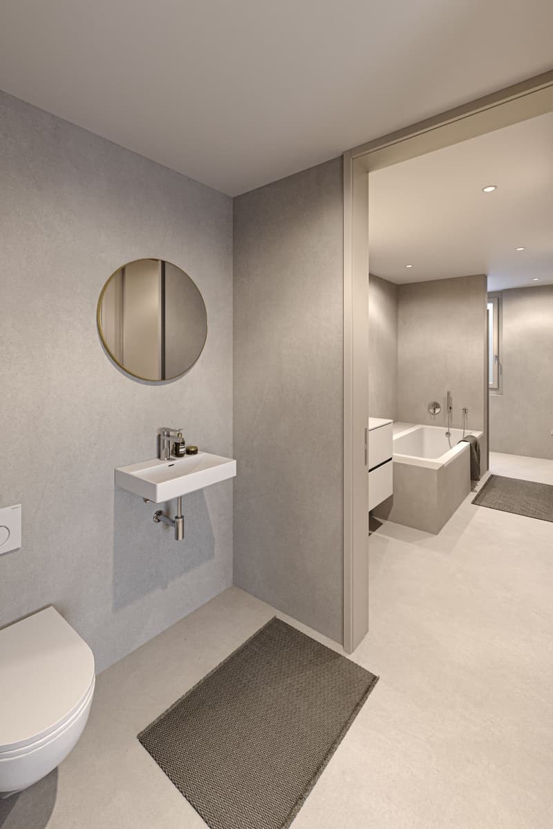 modern bathroom, floor-level bathtub, white porcelain sink, toilet, round mirror, grey wall and floor, open doorway to adjacent room