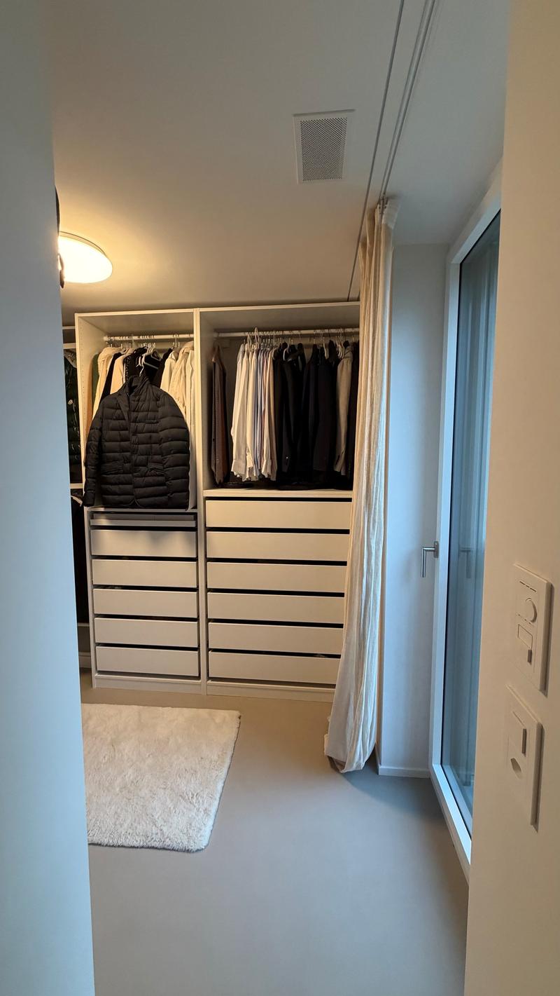 A large walk-in closet with white shelves and drawers, ceiling light, floor rug, and hanging clothes.