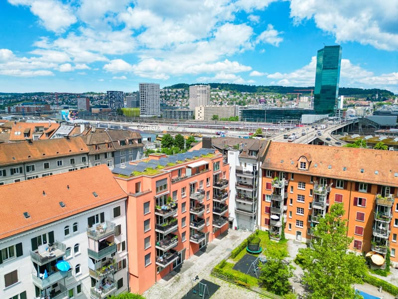 Aerial view of a city skyline with high-rise buildings, residential apartments, and a green park area in the foreground. The buildings have colorful facades and many balconies visible.