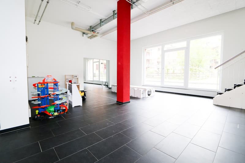 Large open space with white walls, black tile flooring, and a red column. The room contains various toys and equipment, suggesting it may be a commercial or retail space. There are large windows providing natural light.