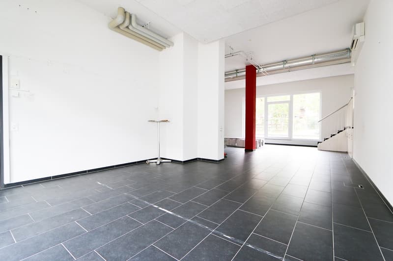 Large open commercial space with white walls, gray tile flooring, and a red column. The space appears to be a retail or office area with a parking place visible.