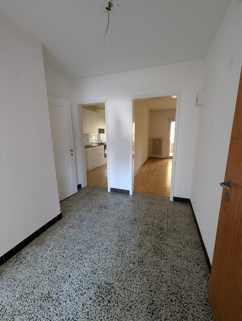 Empty room with white walls, wooden floor, and a door leading to a kitchen
