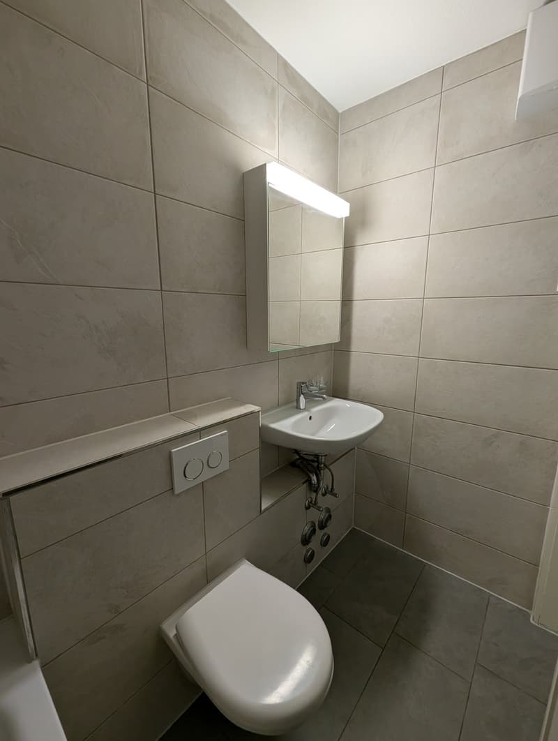 Bathroom with white tiles, white toilet, white sink, mirror, and light above the sink.