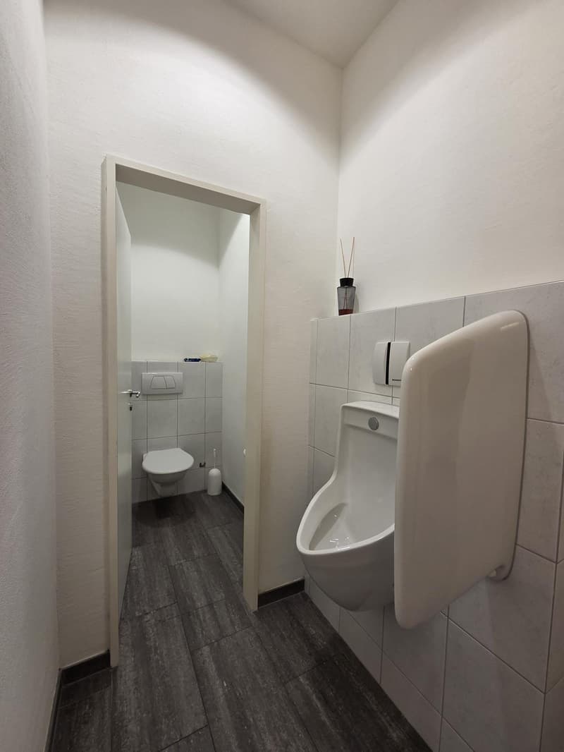 A bathroom with white walls, tiled walls, wooden floor, white toilet, urinal, soap dish, toilet brush, and decorative items