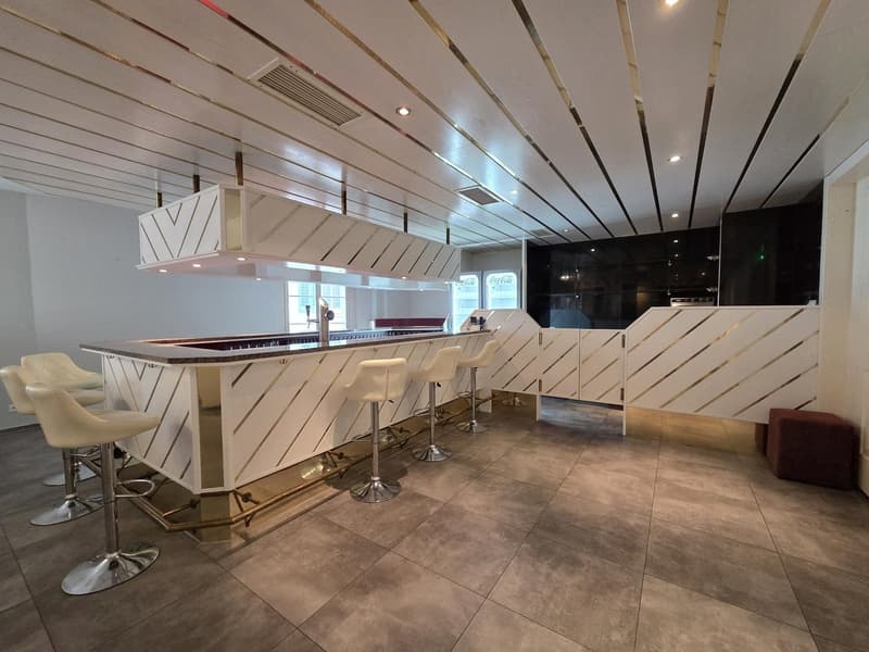 modern bar counter, white leather stools, tiled floor, white and gold accents, ceiling with gold stripes, black cabinets, Coca-Cola fridges