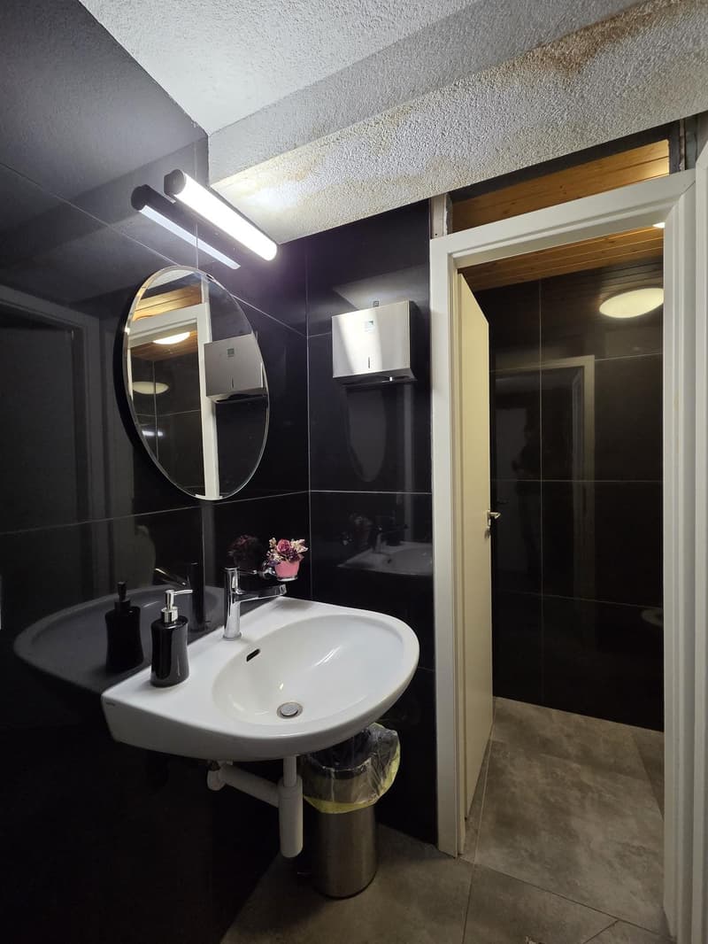 Bathroom with white sink, black tiles, mirror, trash can, soap dispenser, and a door leading to another room