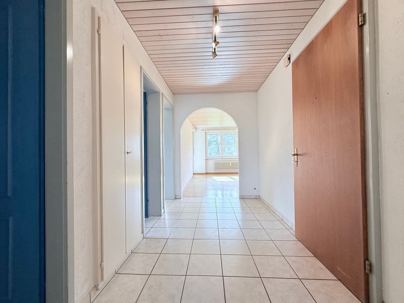 Empty hallway with white tiles, wooden ceiling, white walls, two doors, and an arched doorway leading to a room with a window and radiator.