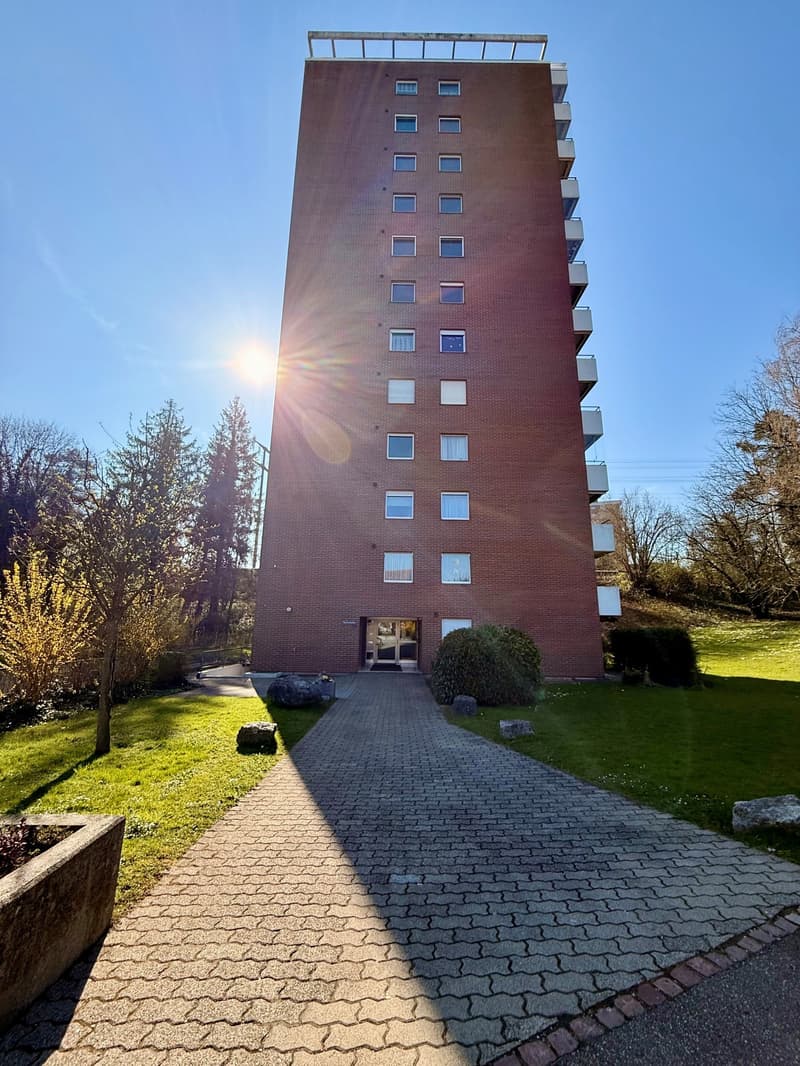 A tall brick building with multiple windows, a paved walkway leading to the entrance, and a grassy area with trees and plants in the background.