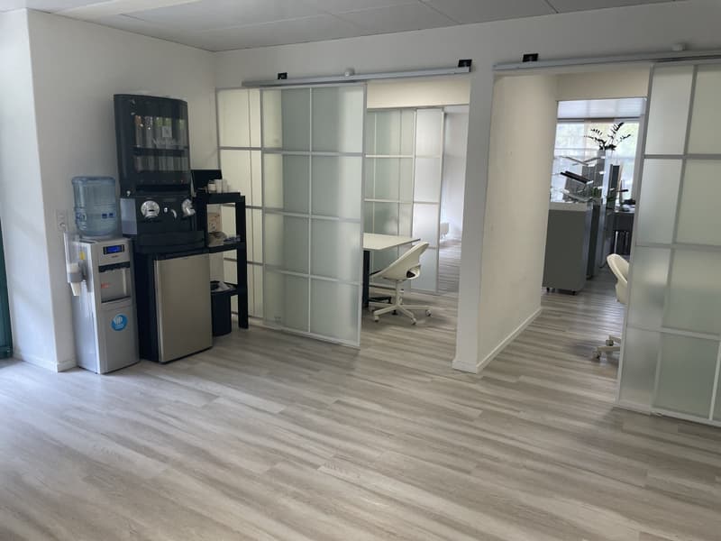 Office space with modern furniture and appliances, hardwood floor, coffee machine and water dispenser on the left side, glass partition doors on the right side.