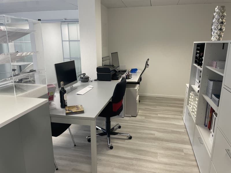 Modern office room with light-colored walls and flooring, two desks with monitors, black office chairs, cabinets with books and a wine bottle on one desk.