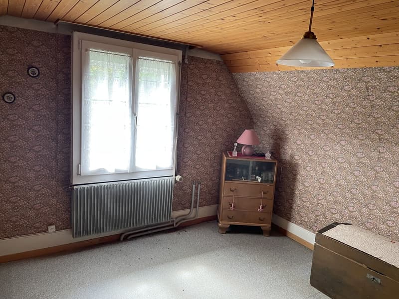 small attic room with wallpaper, large window, radiator, wooden ceiling, lamp, small wooden dresser, footlocker