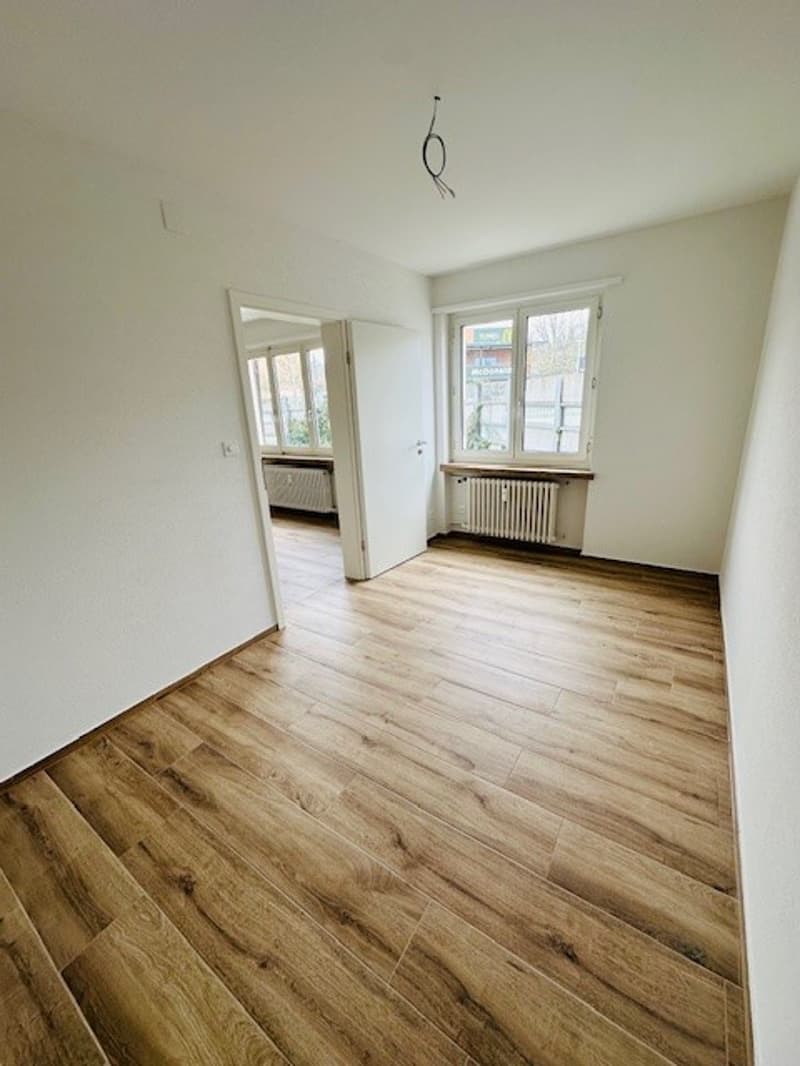 Empty room with wooden floor, white walls, two windows, and radiators.