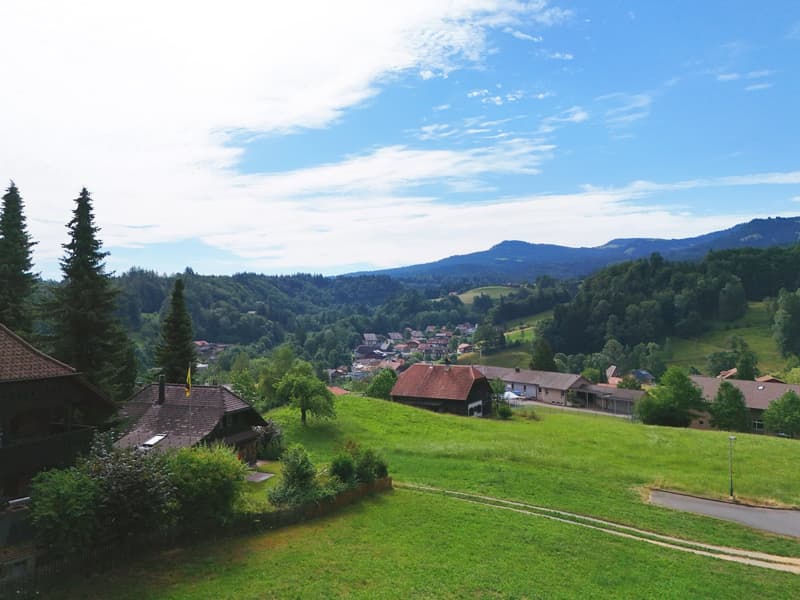 Overlooking the surrounding countryside with a large hill and mountains in the background. Lush green grass, a small road, and several traditional houses with steep roofs.