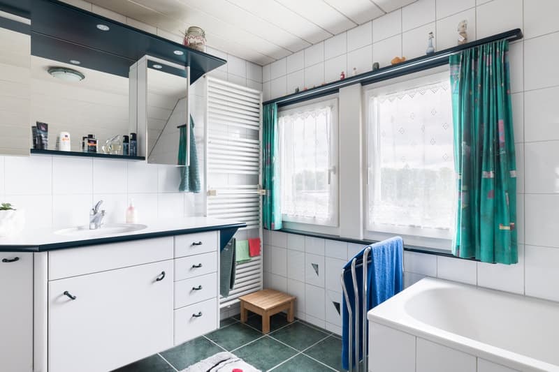Tiled bathroom with white cabinets, black countertops, a washing machine, and a dishwasher. There are curtains on the window and a towel rack with blue towels.