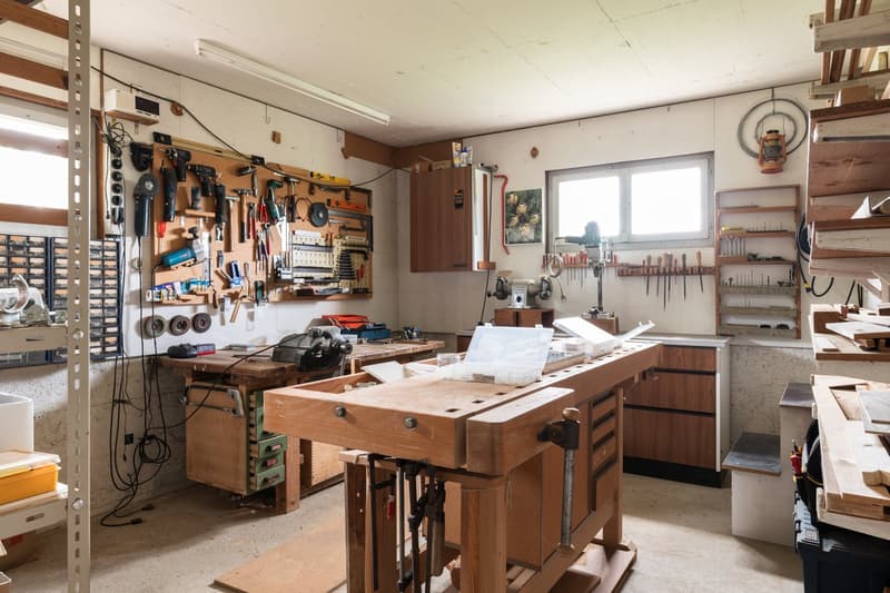 This image shows a well-equipped workshop or garage space. The room is filled with various tools, equipment, and workbenches. The walls are lined with shelves and pegboards displaying a wide range of tools and supplies. There is a large wooden workbench i