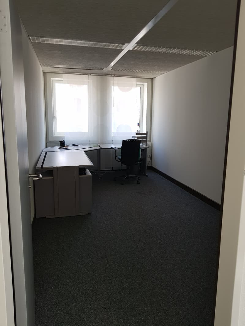 The image shows an office space with a desk, chair, and cabinets. The room has white walls, a dark carpet floor, and a suspended ceiling with a grid pattern. The room appears to be a simple, functional office space.