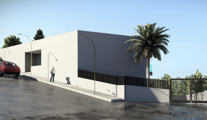 A modern, two-story building with a flat, dark-colored roof and large windows. The building has a minimalist, contemporary design with clean lines and a neutral color palette. There is a palm tree visible in the foreground, and a parking area in front of