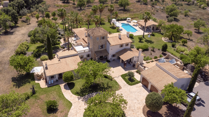 Large two-story Mediterranean-style villa with red tile roof, surrounded by lush landscaping and palm trees. The property includes a swimming pool, garage, and multiple balconies or terraces.