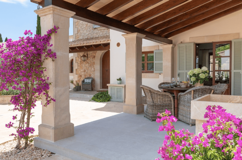 Outdoor patio area with stone columns, wooden beams, and a covered seating area with wicker furniture. Bougainvillea plants in bloom add a vibrant splash of color. The exterior of the house features a stone facade and white walls.