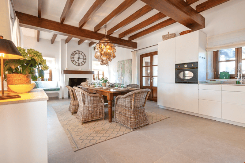 Open-concept kitchen and dining area with exposed wooden beams, a large wooden dining table with wicker chairs, a fireplace, and a modern kitchen with white cabinets and appliances