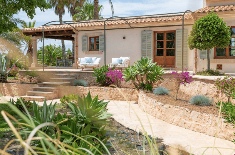Two-story Mediterranean-style house with a tiled roof, white walls, and wooden shutters. The house has a covered patio or terrace with a seating area. The landscaping features lush tropical plants, including palm trees, agave, and flowering shrubs. There