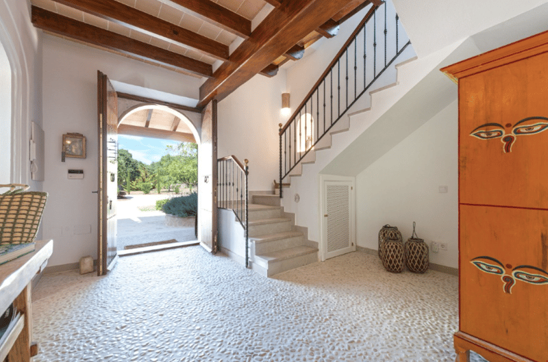 Spacious entryway with wooden beamed ceiling, stone flooring, arched doorway leading to outdoor garden, wooden staircase with metal railing, and decorative wooden furniture pieces