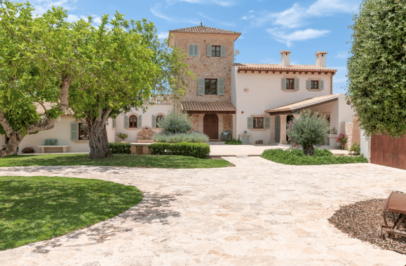 2-story Mediterranean-style villa with terracotta roof tiles, white stucco walls, and large windows. The house is surrounded by a well-maintained garden with trees, shrubs, and a paved driveway leading to a parking area.