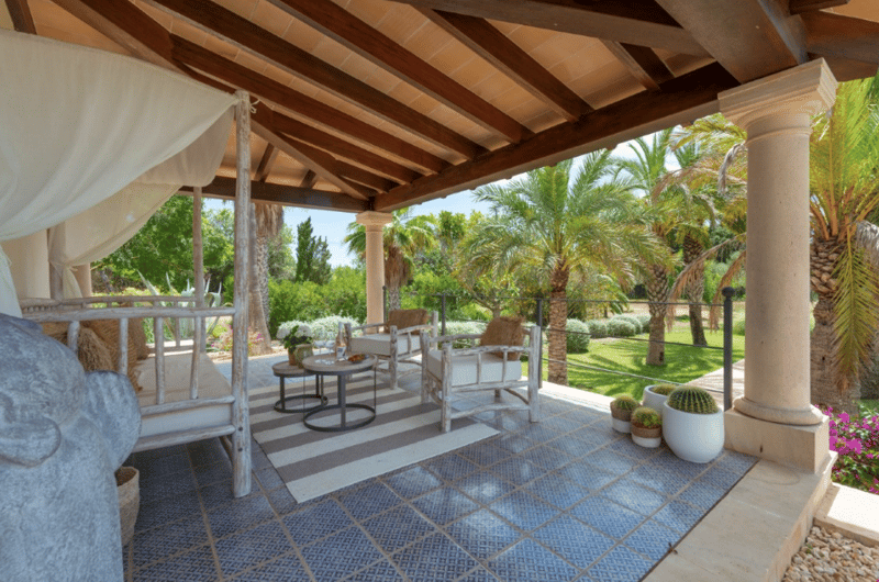 Covered patio with wooden beamed ceiling, white curtains, wicker furniture, coffee table, potted plants, and a view of the lush, palm-filled garden outside