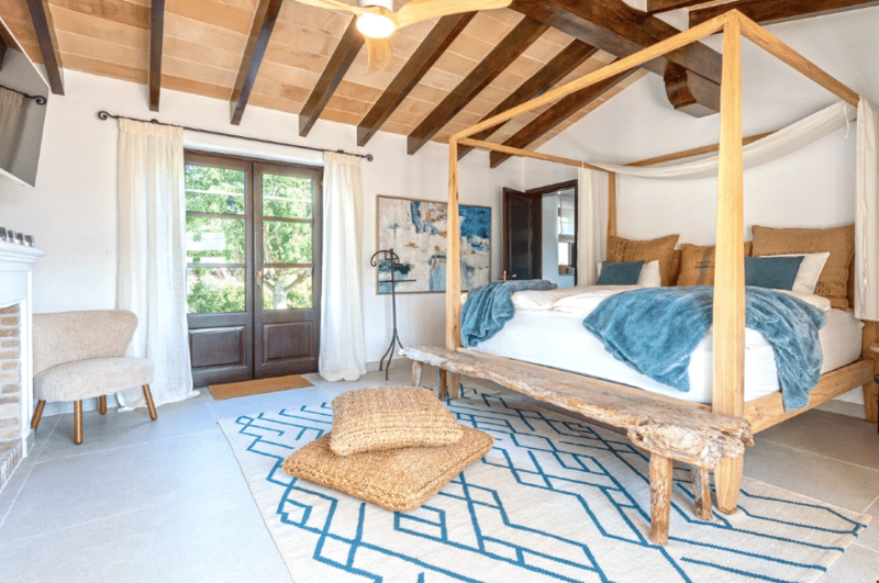 Spacious bedroom with wooden beamed ceiling, large windows, wooden canopy bed, patterned area rug, and rustic wooden furniture