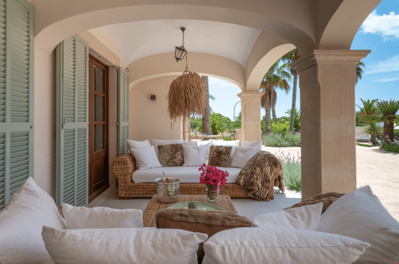 Covered patio with arched entryway, wicker furniture, white cushions, decorative lighting fixture, and view of lush tropical landscaping outside
