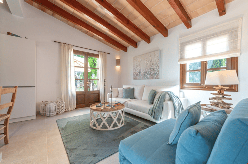 Spacious living room with high wooden ceiling, large windows, white walls, white sofa, blue armchairs, round wooden coffee table, and artwork on the wall