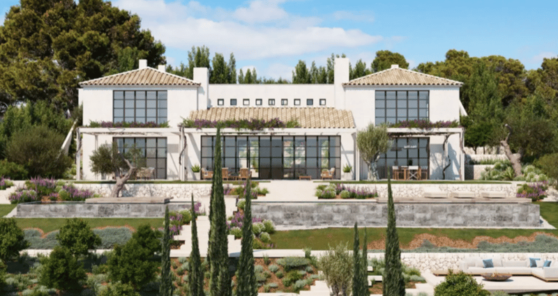 Two-story Mediterranean-style villa with white stucco walls, red tile roof, large windows, and a balcony. The house is surrounded by a lush garden with cypress trees, flowers, and a patio area with outdoor furniture. The property has a scenic view of the