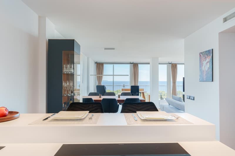 Spacious open-plan living and dining area with large windows offering a scenic view. The room features a dining table with chairs, a built-in cabinet, and a sofa. The overall design is modern and minimalist with a neutral color palette.