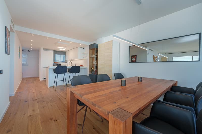 Spacious open-plan living area with a dining table, kitchen, and bar stools. The room features hardwood floors, white walls, and large windows. The kitchen has modern appliances including a dishwasher. The dining table is made of wood with a unique design