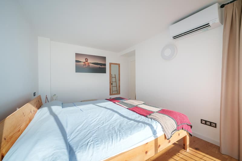 Spacious bedroom with white walls, wooden bed frame, large window, and air conditioning unit. The room features a colorful patterned blanket on the bed and a framed artwork on the wall.