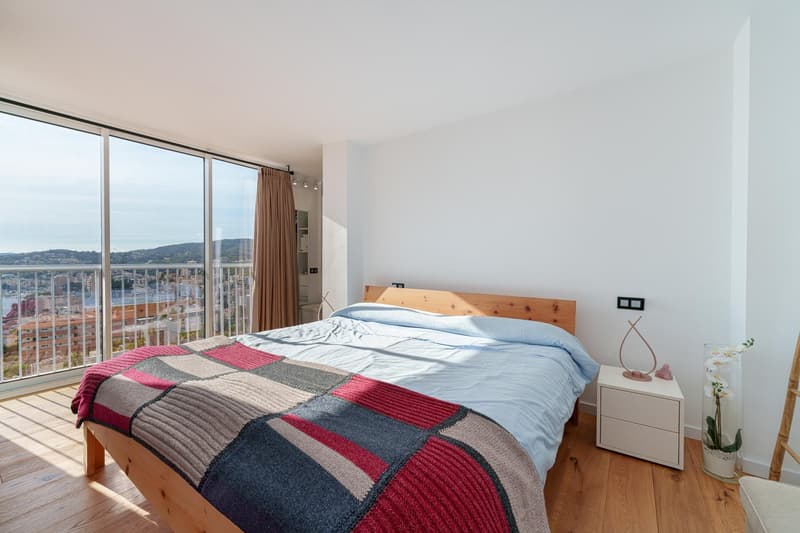Spacious bedroom with large windows offering a panoramic view of the city, wooden bed frame, patterned bedding, and a bedside table with a decorative vase.