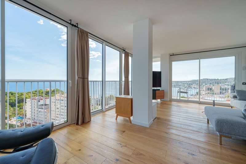 Spacious living room with large windows offering a panoramic view of the city and harbor. The room features hardwood flooring, a TV stand, and a couch. Curtains are drawn over the sliding glass doors leading to a balcony or terrace.