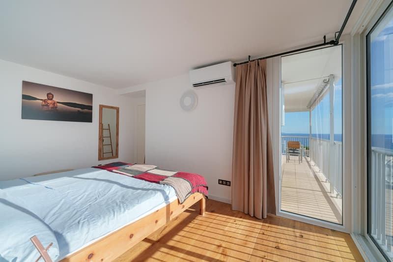 Spacious bedroom with large windows and sliding glass door leading to a balcony with a view of the ocean. The room features a wooden bed frame, a wall-mounted air conditioning unit, and a framed artwork depicting a Buddha statue. The floor is made of wood