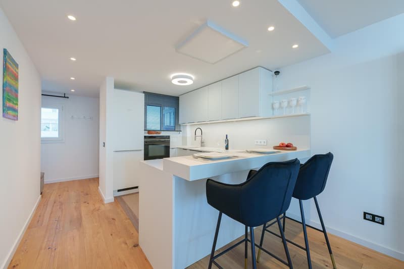 Modern, open-concept kitchen with white cabinets, quartz countertops, stainless steel appliances including a dishwasher, and navy blue bar stools. The kitchen has a clean, minimalist design with recessed lighting and hardwood flooring.