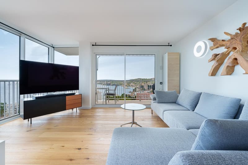Spacious living room with large windows offering a scenic view, hardwood flooring, gray sectional sofa, coffee table, and a wall-mounted TV cabinet