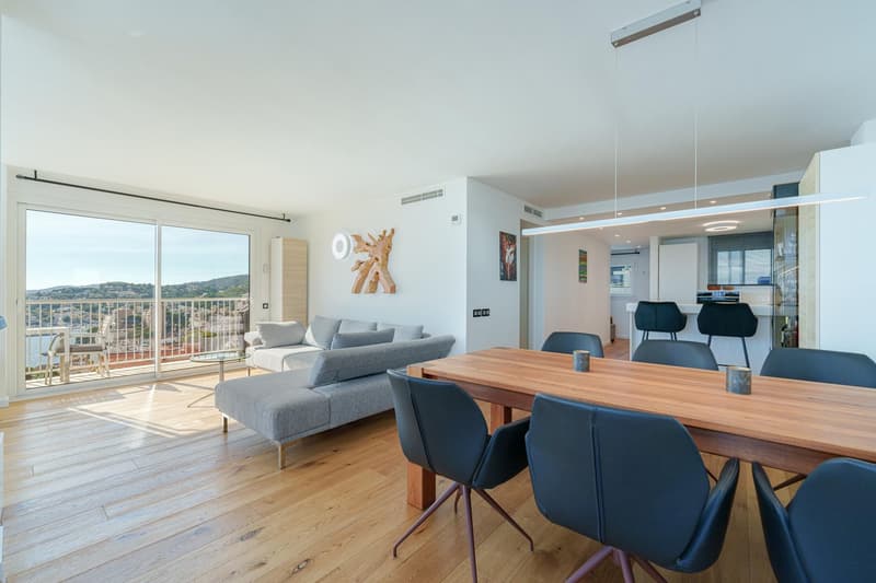 Spacious open-plan living and dining area with large windows providing a panoramic view. The living room features a comfortable grey sofa set, while the dining area has a wooden table and black chairs. The room has a modern and minimalist design with wood