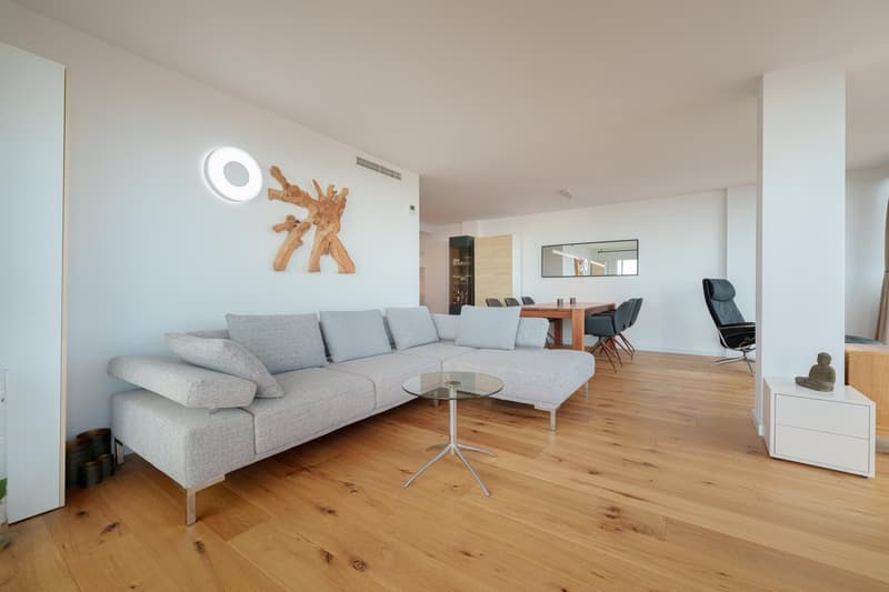 Spacious living room with a large gray sectional sofa, glass coffee table, and wooden dining table with chairs. The room has hardwood floors, white walls, and a decorative wooden sculpture on the wall. There is also a balcony or terrace visible through th