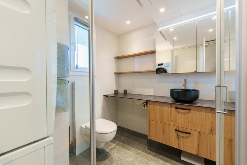 Modern bathroom with wooden vanity, black vessel sink, shelves, and a washing machine and dishwasher installed. The bathroom has a glass shower enclosure and tile flooring.