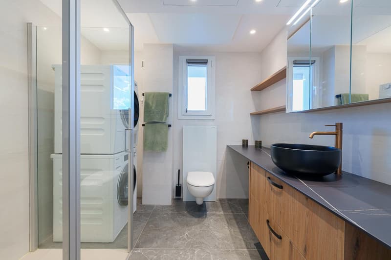 Modern bathroom with a vanity unit, black vessel sink, and a washing machine. The room has a tiled floor and a window for natural light. There is also a toilet and shelving for storage.
