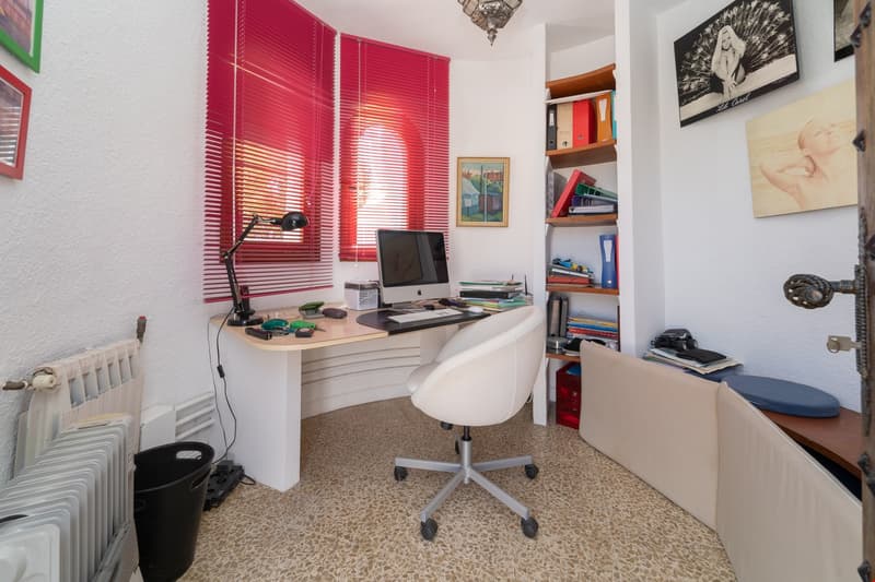 Spacious home office with a white desk, computer, and various office supplies. The room has red blinds covering the windows and shelves with books and decor. The floor is tiled with a patterned design.
